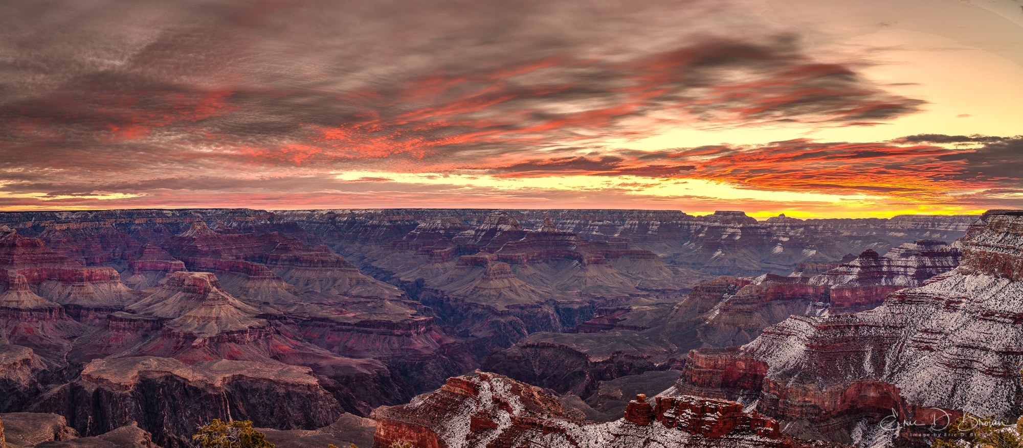 Panoramic sunrise over the Grand Canyon from the south rim, a heavy cloud deck lit red from underneath with a gold band on the horizon, snow on the far rim and on the foreground rock.