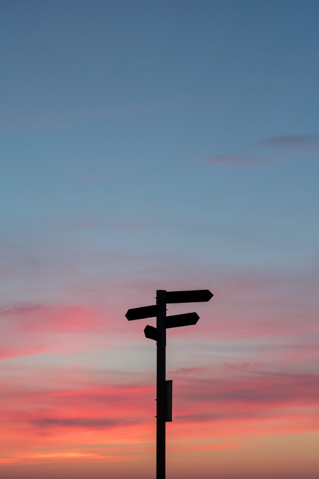 Silhouette of a directional signpost against a pink and blue sunset sky