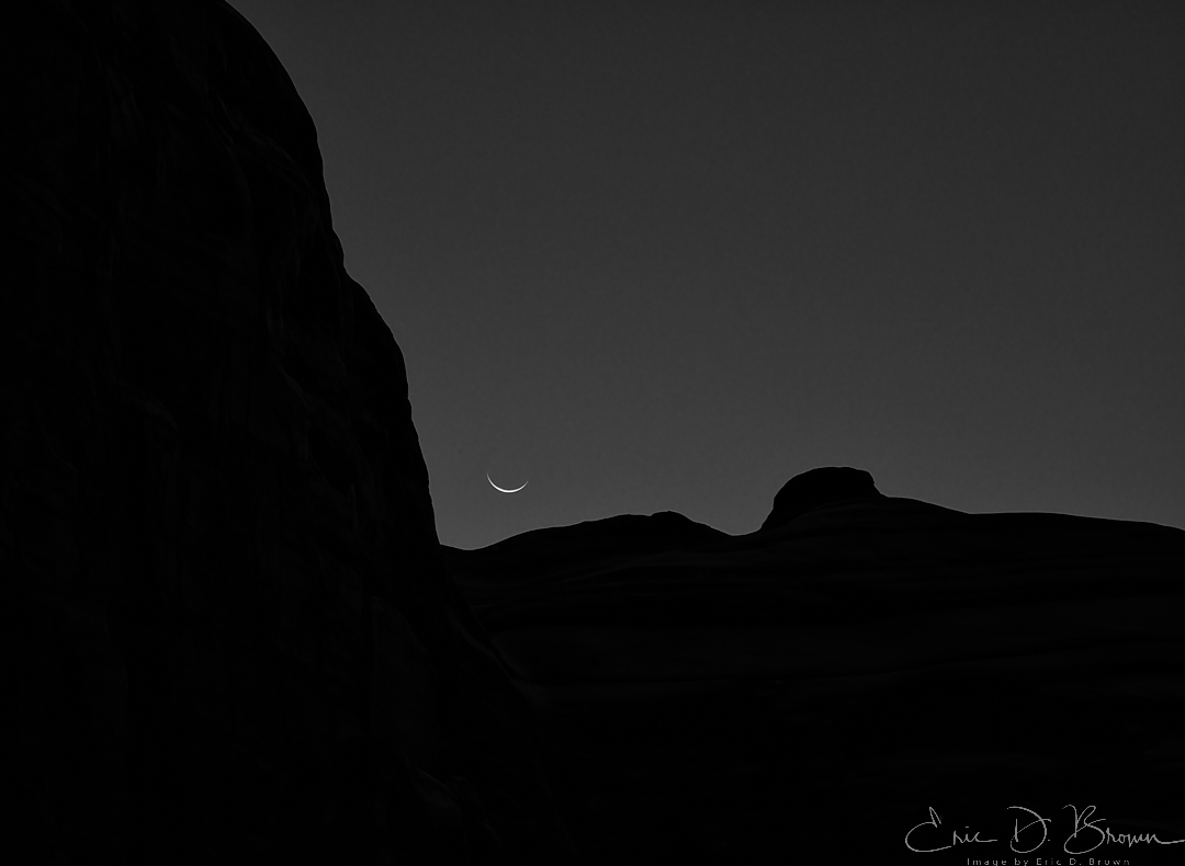 A thin sliver of moon low over the rock walls at the entrance to Park Avenue in Arches National Park, Utah, converted to black and white against a plain sky