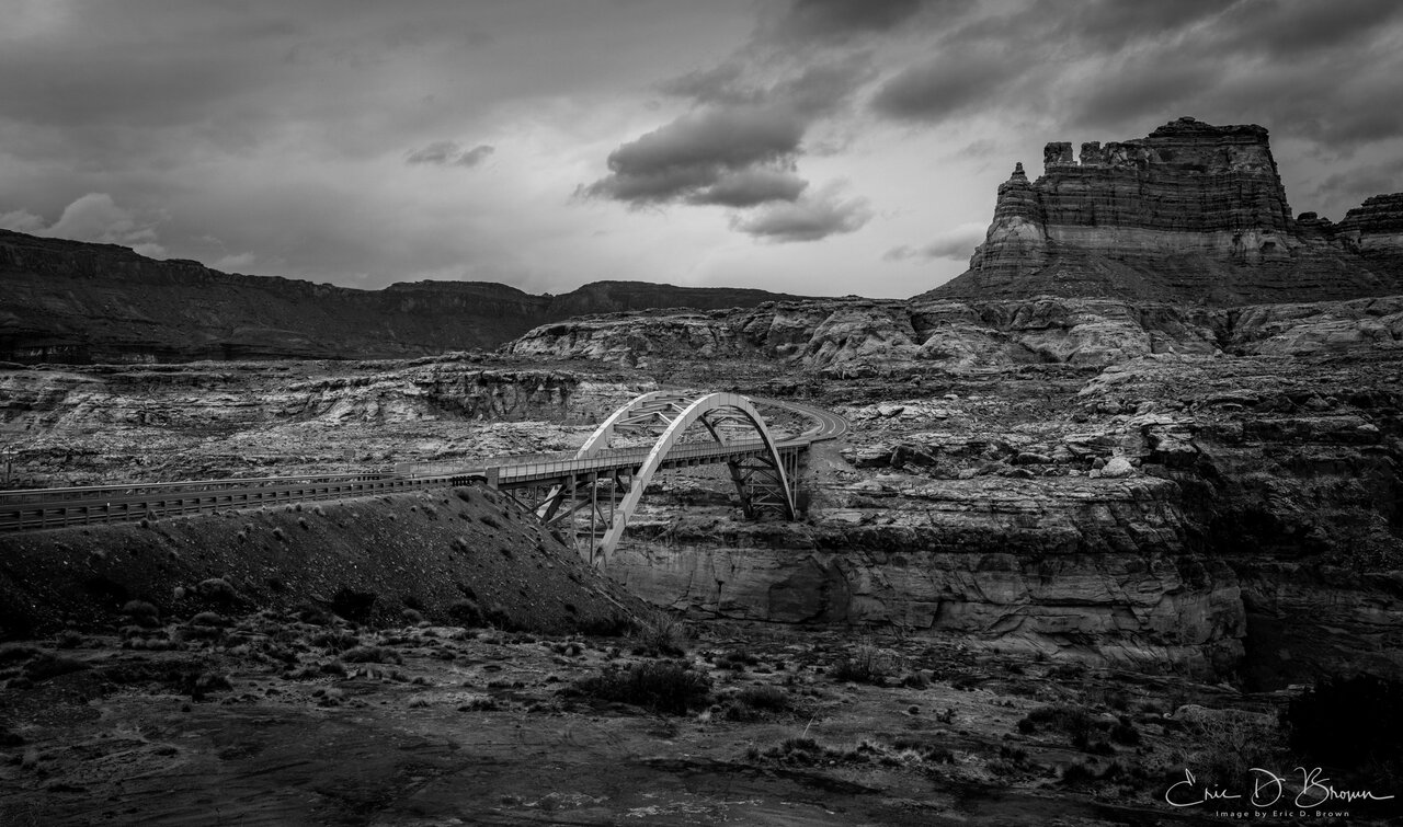 A steel arch bridge carrying a highway over a deep canyon of the Colorado River in southern Utah, beneath a heavy storm sky, in black and white.