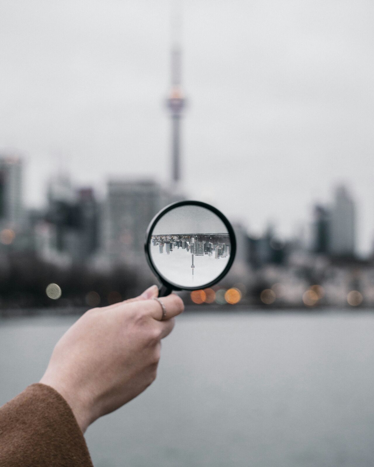 A person holding a magnifying glass up to the daytime sky, bringing the distant CN Tower in Toronto into sharp focus through the lens