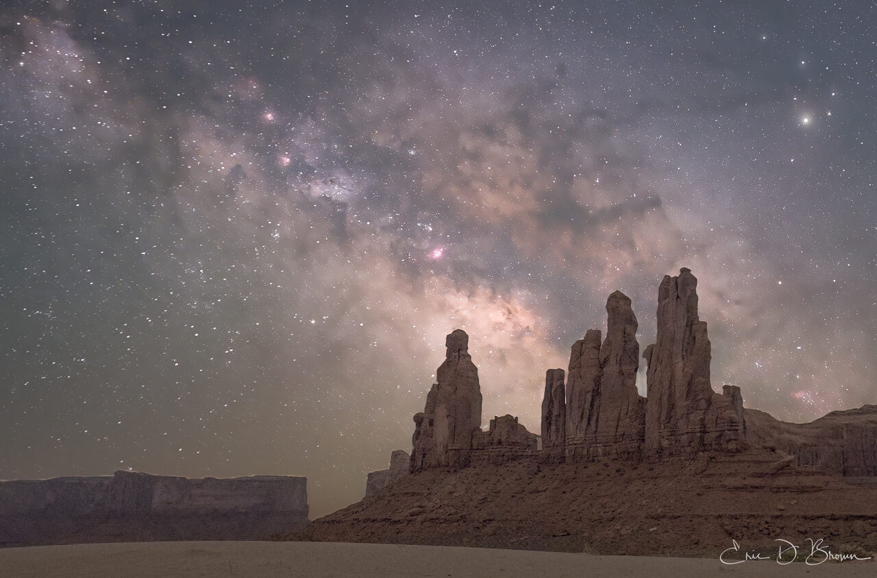 The Totem Pole and nearby spires of Monument Valley silhouetted against the Milky Way core rising pink and gold behind them.
